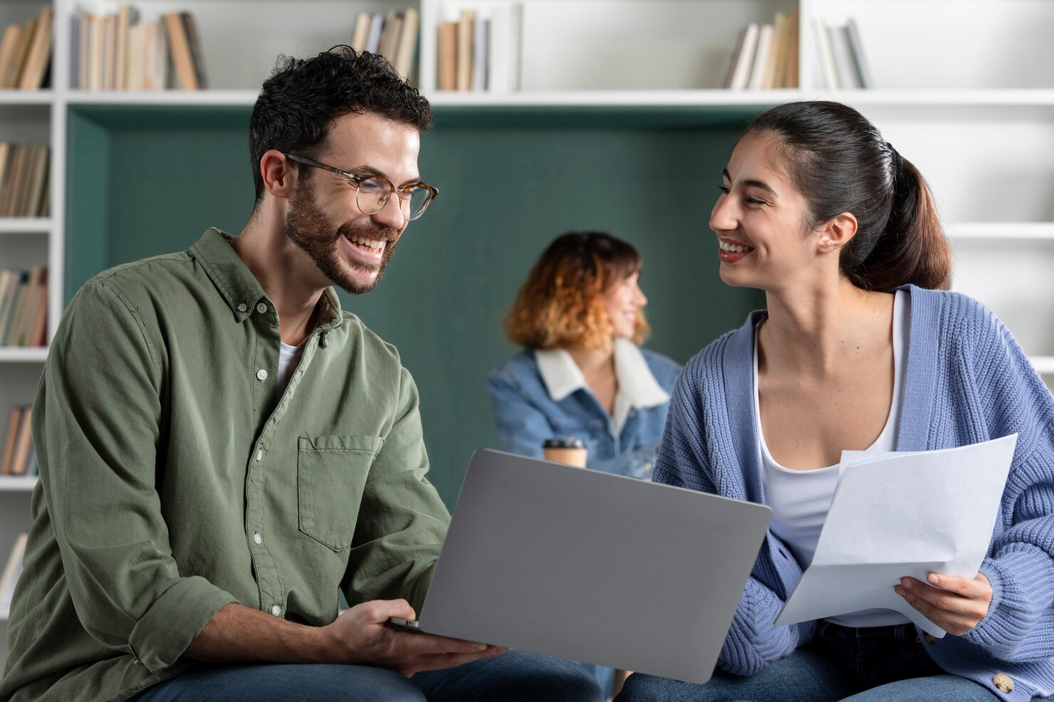 A young man, left, and young woman, right, smile as the man shares his laptop screen while the woman holds printed materials.